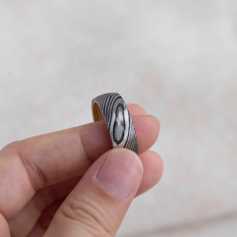 Black and silver damascus steel ring with a wood inlay, held between fingers.