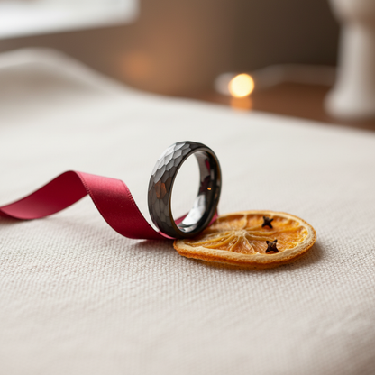 A black faceted ring rests on a dried orange slice, accompanied by a red ribbon, creating a cozy, festive scene.