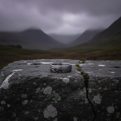 Bevelled 8mm artisan crucible ring in dark tones by Forge and Lumber, displayed on a stone surface outdoors.