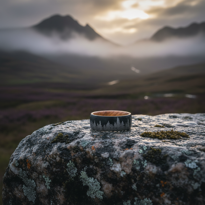 A detailed ring with forest engraving resting on a mossy stone, set against a misty mountain landscape at sunset.