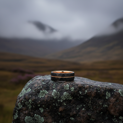 Stylish black and copper ring resting on a moss-covered rock, with a misty mountain landscape in the background.